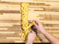 Hands braiding strands of dough for Christmas Vánočka (Houska) on a wooden board.