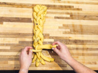 Hands braiding strands of yellow dough studded with dark bits for Christmas Vánočka (Houska) on a wooden board.