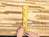 Hands braiding strands of yellow dough for Christmas Vánočka (Houska) on a wooden board.