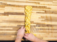 Hands braiding strands of Christmas Vánočka (Houska) dough studded with raisins on a wooden counter.