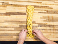 Hands braiding pale dough strands into Christmas Vánočka (Houska) on a wooden board