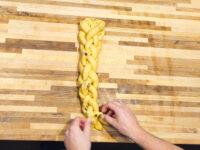 Hands braiding a strand of raisin-studded dough for Christmas Vánočka (Houska) on a wooden board.