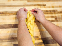 Hands braiding yellow dough strands for Christmas Vánočka (Houska) on a wooden board.