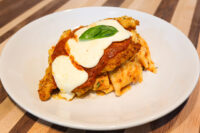 Chicken Parmesan cutlet topped with marinara, melted mozzarella, and a basil leaf, served over wide pasta ribbons on a white plate