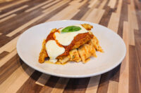 Chicken Parmesan cutlet topped with marinara and melted mozzarella, garnished with basil, served over pasta on a white plate.