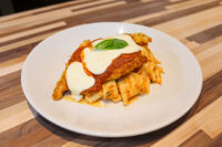 Chicken Parmesan topped with marinara sauce, melted mozzarella, and a basil leaf, served over tube-shaped pasta on a white plate.