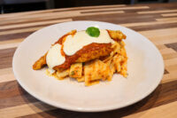 Chicken Parmesan cutlet topped with tomato sauce, melted cheese, and a basil leaf, served over pasta on a white plate.