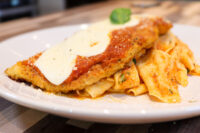 Breaded Chicken Parmesan topped with marinara and melted cheese, served beside ribbon pasta on a white plate.