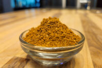 Small glass bowl mounded with reddish-brown Garam Masala Seasoning on a wooden table.
