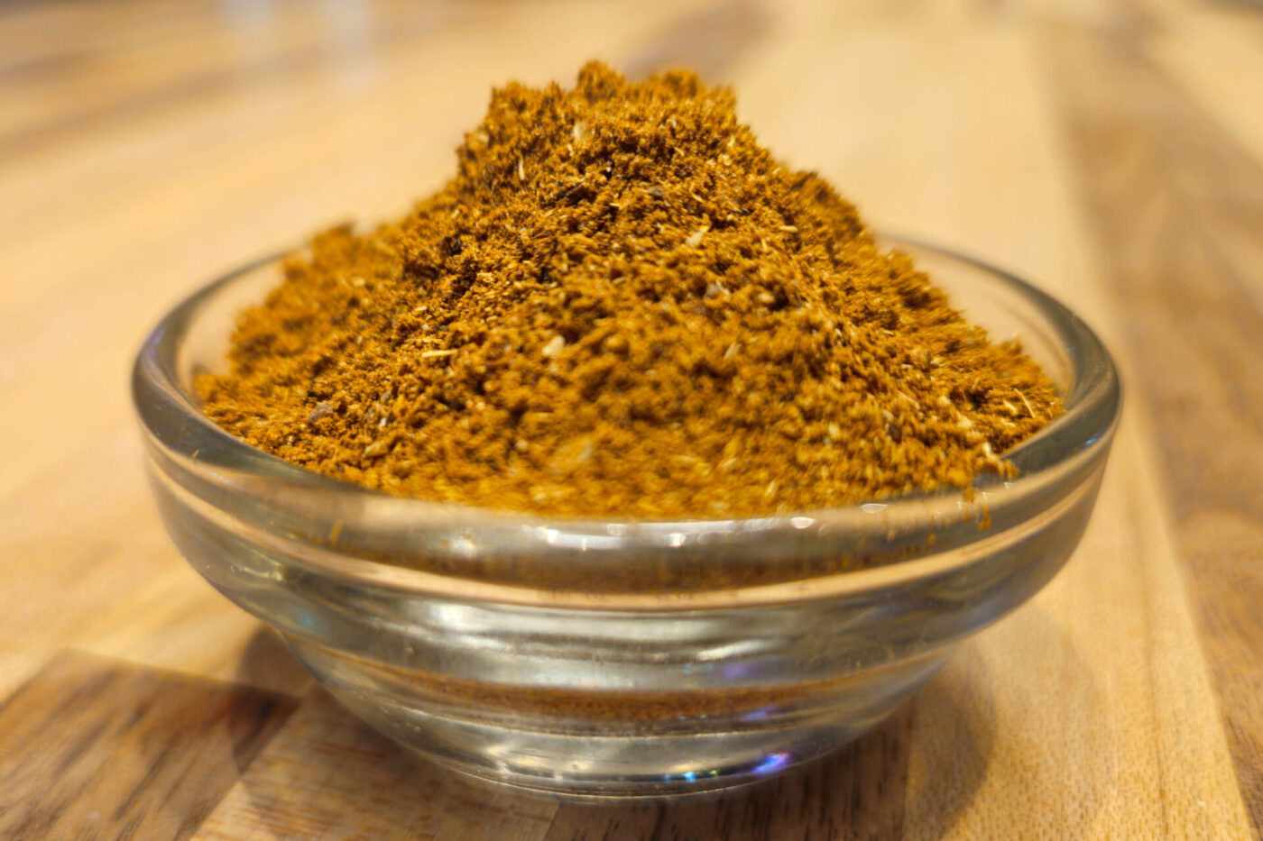 Small glass bowl heaped with reddish-brown Garam Masala Seasoning powder on a wooden countertop.