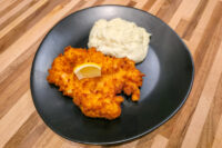 Cheese Battered Chicken Schnitzel Kuře v Sýrovem Těstíčku with lemon wedge and mashed potatoes on a dark plate.