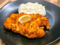 Cheese Battered Chicken Schnitzel Kuře v Sýrovem Těstíčku with a lemon wedge and mashed potatoes on a dark plate.
