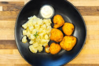 Four pieces of breaded Fried Cauliflower Smažený Květák on a black plate with diced potatoes and dip.
