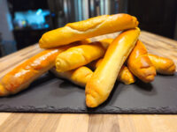 Golden-brown Czech Rolls Rohliky, elongated and tapered, piled on a dark slate board on wood.