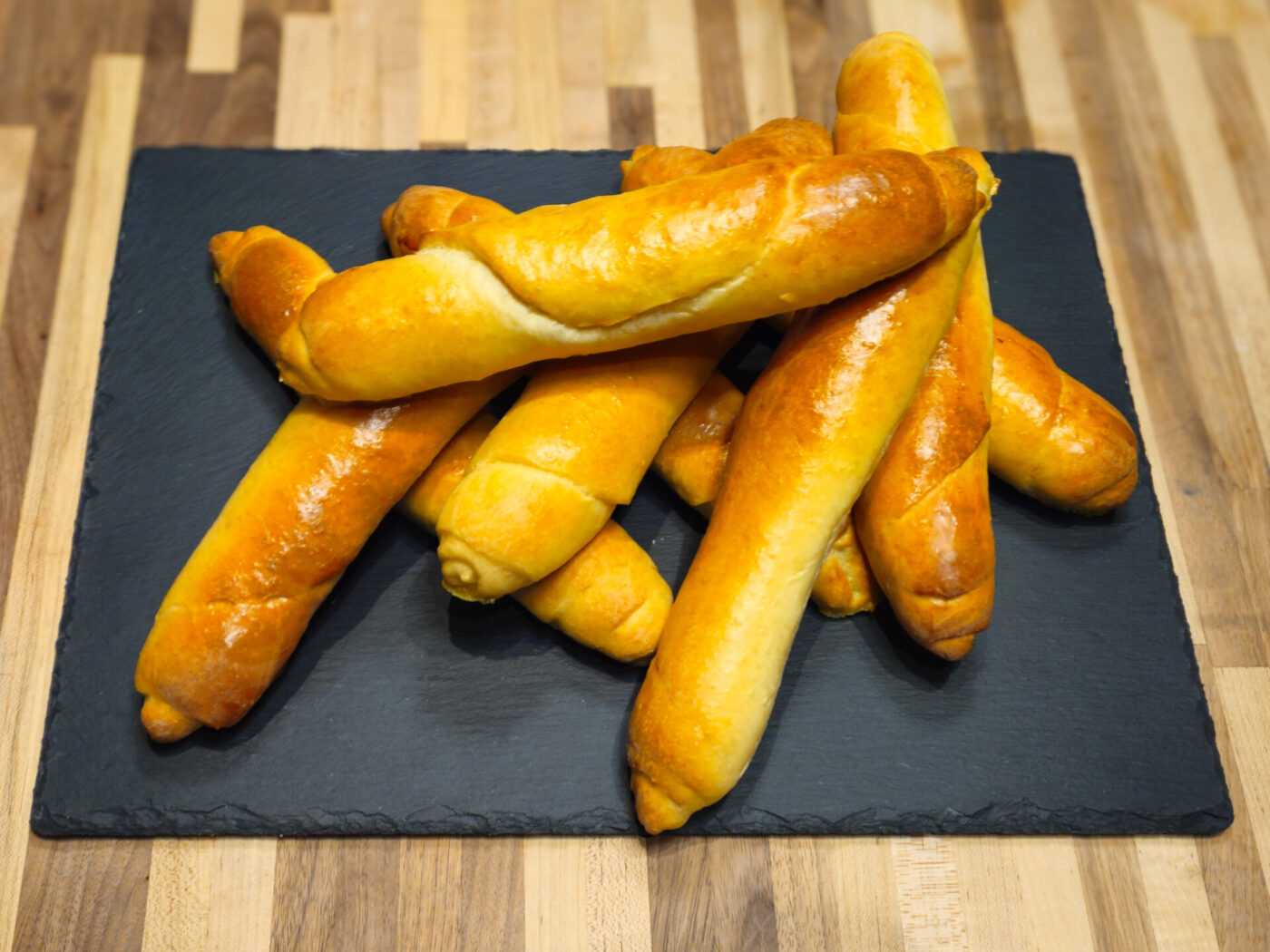 Pile of glossy golden Czech Rolls Rohliky, shaped into long tapered sticks, on a dark slate board.