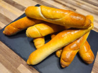 A pile of golden-brown Czech Rolls Rohliky with scored tops, stacked on a dark slate board.