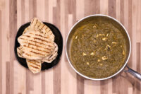 Grilled Naan Bread pieces stacked on a dark plate next to a pan of green curry with pale cheese cubes, on a wooden countertop.