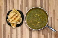 Pan of green Spinach Palak Paneer curry beside grilled flatbread wedges on a black plate.