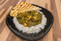 Spinach Palak Paneer with cubed paneer over rice, served with grilled flatbread on a black plate.