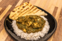 Spinach Palak Paneer with cubed paneer over rice, served with grilled flatbread on a black plate.