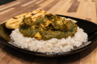 Black plate of Spinach Palak Paneer over white rice with grilled naan bread on the side.