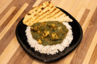 Spinach Palak Paneer curry with cubed paneer over rice, served with grilled naan on a black plate.