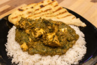 Spinach Palak Paneer with paneer cubes over rice, served with grilled naan on a black plate.