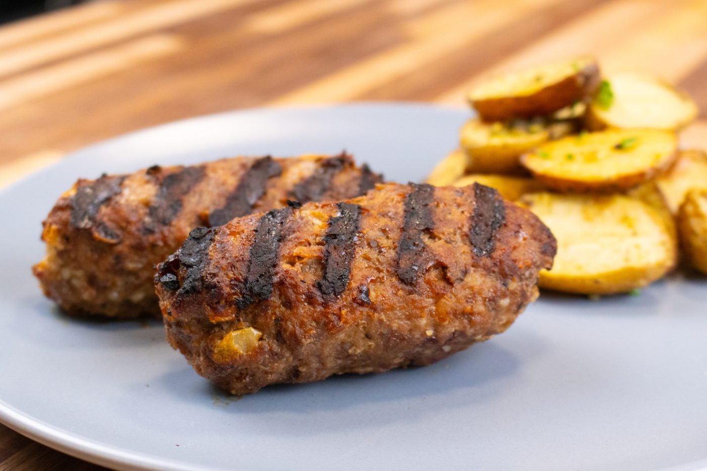 Two grilled Čevapčiči Sausages With Cheese with char marks, served with roasted potato slices on a grey plate.