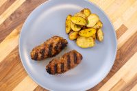 Two grilled Čevapčiči sausages with grill marks beside roasted sliced potatoes on a grey plate.