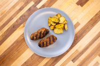 Two grilled Čevapčiči sausages with char marks beside roasted potato wedges on a grey plate.