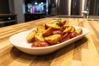 Baby Red Potatoes with Butter, quartered and glazed with a buttery sheen, served in a white oval dish on a wooden counter with a kitchen blurred behind it.