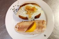 Baked Potato with Herb Sour Cream, topped with sautéed onion strands, plated on a leaf-patterned dish beside a poached fish fillet and lemon wedge on a steel counter.