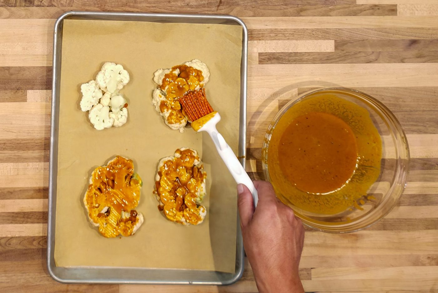 Hand brushes orange Buffalo sauce onto cauliflower pieces for Buffalo Cauliflower Steak on a baking sheet.