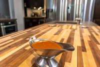 A metal gravy boat filled with glossy orange-red Buffalo Sauce sits on a striped wooden countertop.