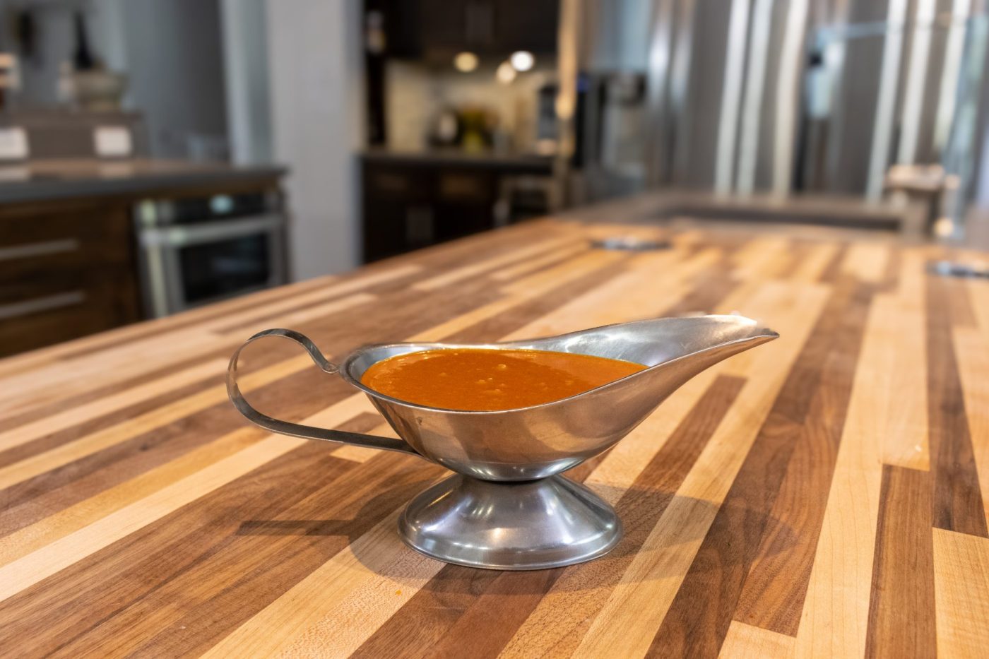 Orange-red Buffalo Sauce served in a metal gravy boat on a striped wooden countertop.