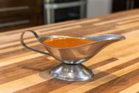 Buffalo Sauce pooled in a stainless steel gravy boat on a wooden countertop.