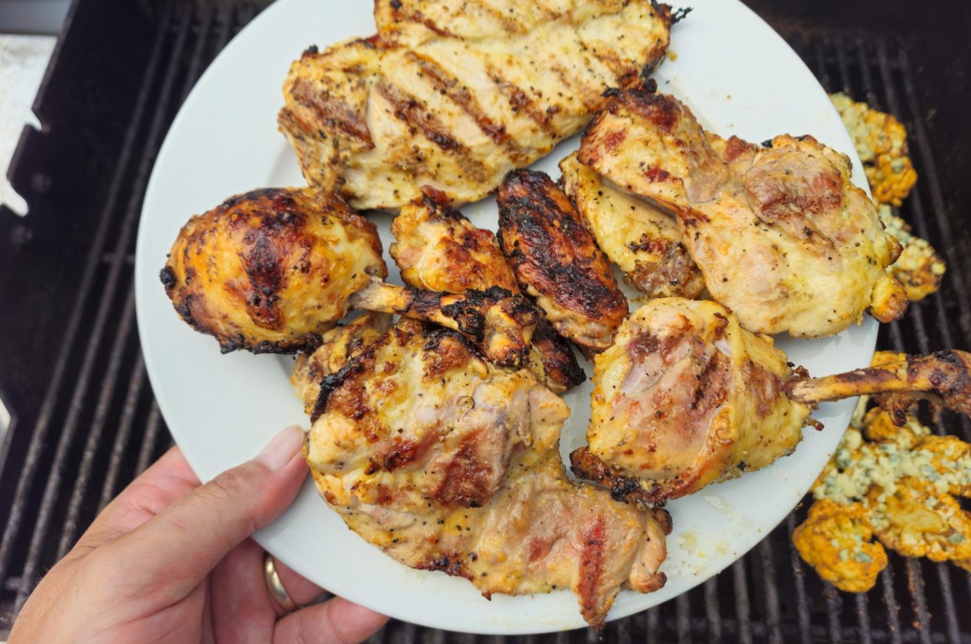 Hand holding a white plate of grilled Lemon and Garlic Chicken pieces over a barbecue grill.
