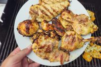 Hand holding a white plate of grilled Lemon and Garlic Chicken pieces over a barbecue grill.