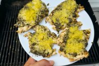 Four Pesto Chicken Breasts topped with green pesto and yellow parmesan on a white plate near a grill.