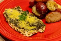 Pesto Chicken Breasts topped with green pesto and golden parmesan, parsley garnish, beside roasted red potatoes.