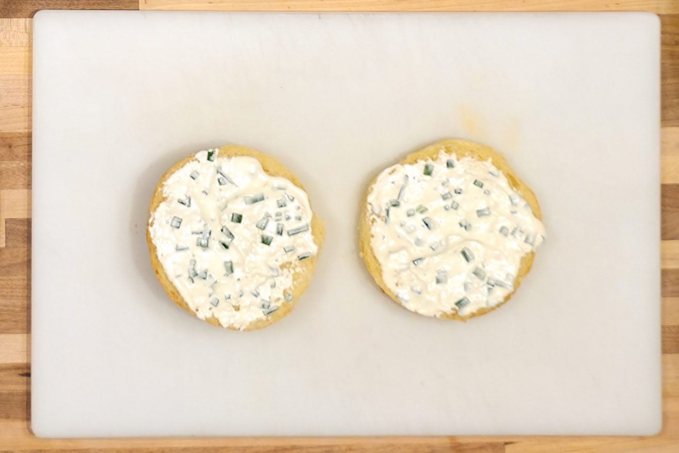 Two bun halves spread with a creamy white sauce and chopped green herbs, resting on a white cutting board as part of a Pesto Chicken Sandwich with Parmesan