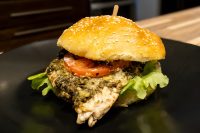 Pesto Chicken Sandwich with Parmesan on a sesame bun with tomato and lettuce, skewered on a dark plate.