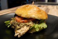 Pesto Chicken Sandwich with Parmesan on a sesame bun with tomato and arugula, on a dark plate.
