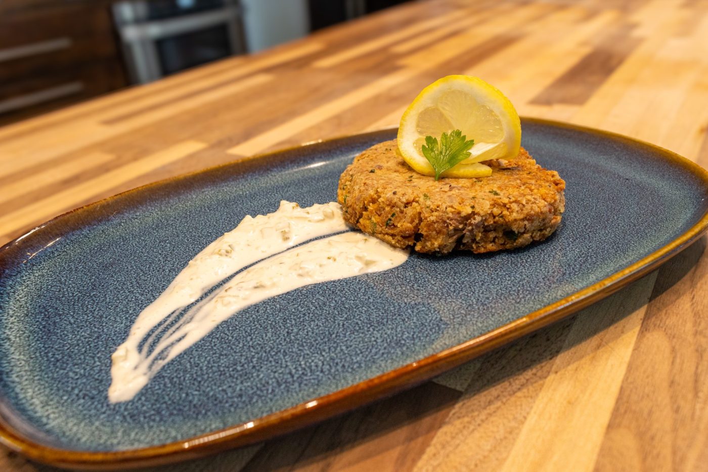 Tuna Cakes Golden breaded Tuna Cake topped with a lemon slice and herb sprig on a blue plate with sauce swipe.