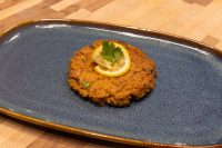 Tuna Cakes A golden-brown Tuna Cake topped with a lemon slice and herb sprig on a dark blue oval plate