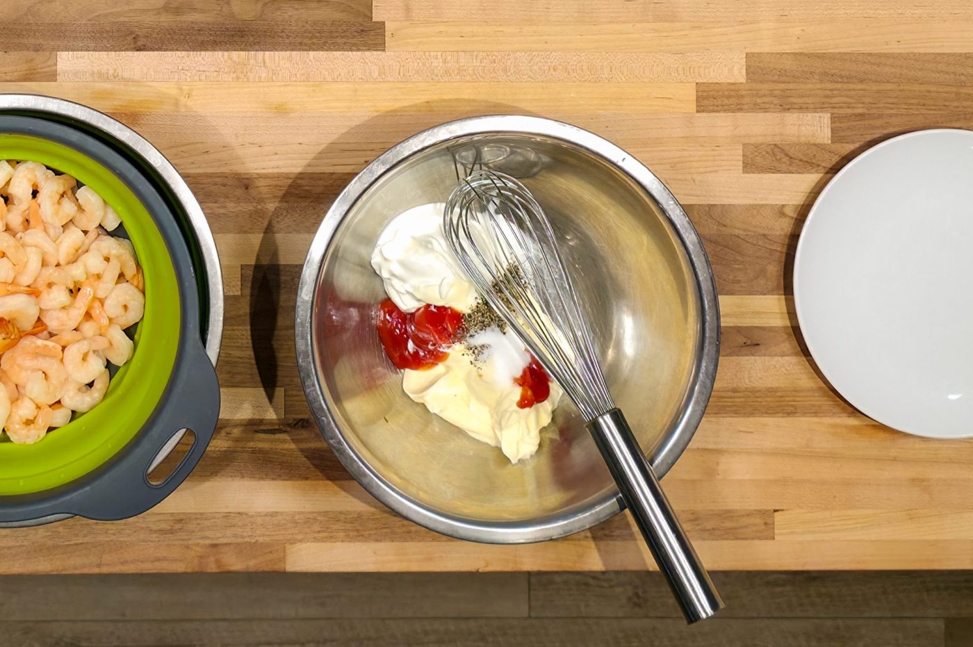 Metal bowl with mayonnaise, ketchup, and whisk beside shrimp and an empty plate for Shrimp Cocktail.