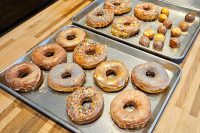 Two trays of glazed Donuts and Donut Holes, some topped with sprinkles, on a wooden counter