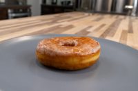 A glazed ring donut from Donuts and Donut Holes sits on a dark gray plate on a wooden counter.