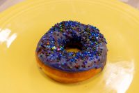 A single Donuts and Donut Holes ring with purple glaze and colorful sprinkles on a yellow plate.