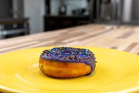 A single Donuts and Donut Holes pastry with purple glaze and rainbow sprinkles on a yellow plate.